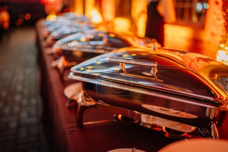 Riga, Latvia - February 16, 2024 - a row of shiny chafing dishes on a buffet table, reflecting the warm, ambient light of the venue.