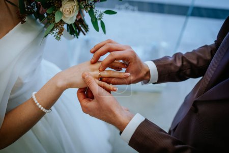 Riga, Lettland - 20. Januar 2024 - Ein Bräutigam legt der Braut einen Ehering an den Finger, beide formell gekleidet, mit einem Brautstrauß im Blick.