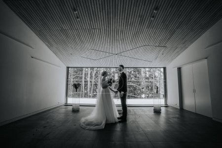 Riga, Lettland - 20. Januar 2024 - Braut und Bräutigam stehen einander gegenüber, Händchen haltend, in einem modernen Raum mit großen Fenstern mit schneebedeckten Bäumen draußen