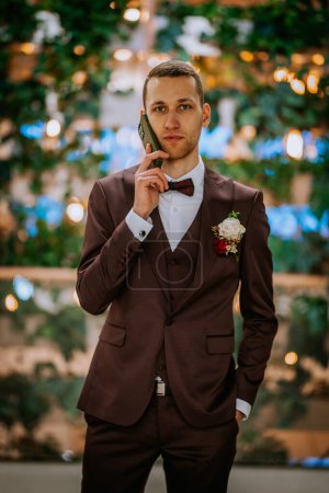 Riga, Lettonie - 20 janvier 2024 - Un marié en costume marron est au téléphone, avec une boutonnière sur son revers et un fond bokeh de lumières et de feuilles.