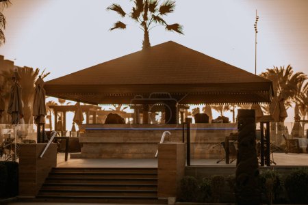 Agadir, Maroc - 25 février 2024 - un belvédère au bord de la piscine avec un toit brun au coucher du soleil. Deux parapluies fermés et des palmiers sont visibles dans une lumière chaude et dorée