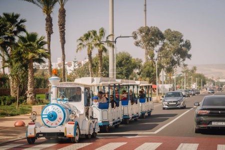 Téléchargez les photos : Agadir, Maroc - 25 février 2024 - Train routier touristique décoré avec passagers, palmiers, ciel bleu, milieu urbain, croisement, véhicules. - en image libre de droit