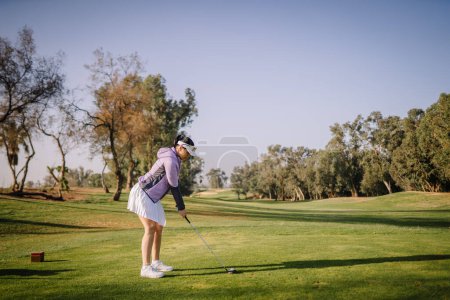 Agadir, Morocco - February 25, 2024 - A female golfer in a white skirt and purple top is in mid-swing on a golf course.