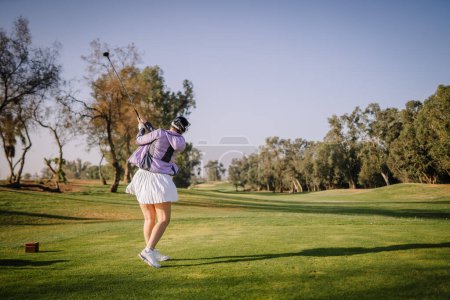 Agadir, Marokko - 25. Februar 2024 - Eine Golferin in lila Top und weißem Rock beendet ihren Schwung auf einem sonnigen Golfplatz.