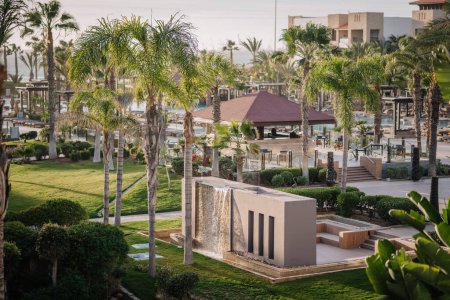 Agadir, Morocco - February 25, 2024 - Resort garden with palm trees, waterfall feature, grass lawn, and poolside gazebo at dusk