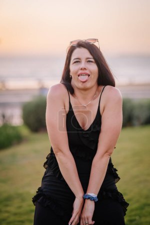 Téléchargez les photos : Une femme ludique qui sort sa langue, portant une robe noire à volants et des lunettes de soleil sur la tête, avec une plage et un coucher de soleil en arrière-plan. - en image libre de droit