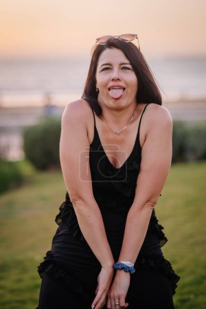 Photo for A playful woman sticking out her tongue, wearing a black ruffled dress and sunglasses on her head, with a beach and sunset in the background. - Royalty Free Image