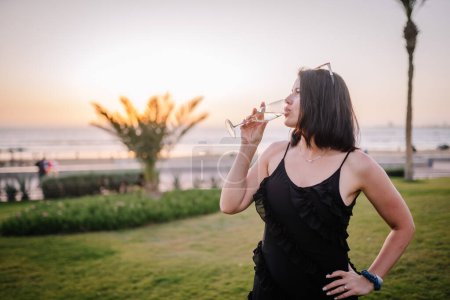 A woman in a black dress sips champagne by the seaside at sunset, with palm trees in the background and sunglasses atop her head