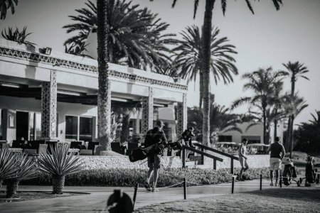 Téléchargez les photos : Agadir, Maroc - 28 février 2024- Golfeurs avec équipement marchant devant un club-house encadré de palmiers, sur une photo en noir et blanc avec une sensation vintage - en image libre de droit