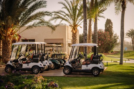 Téléchargez les photos : Agadir, Maroc - 28 février 2024- Chariots de golf garés sur un sentier sur un terrain de golf avec des palmiers et un bâtiment en arrière-plan, baignés de soleil chaud - en image libre de droit