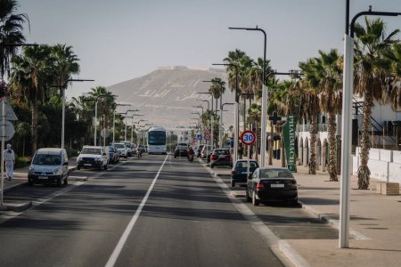 Téléchargez les photos : Agadir, Maroc - 28 février 2024- Une rue urbaine avec des palmiers et des voitures, un bus au loin, des panneaux de signalisation, un ciel dégagé et une montagne en arrière-plan - en image libre de droit