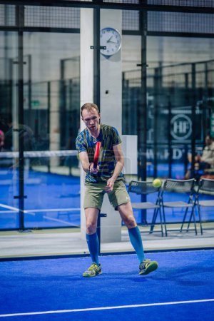 Foto de Riga, Letonia - 3 de marzo de 2024 - Un jugador está listo para atacar en un juego de pádel, usando una camisa a rayas, pantalones cortos caqui y calcetines azules de rodilla en una cancha azul interior.. - Imagen libre de derechos