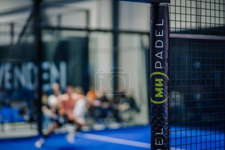 Photo for Riga, Latvia - March 3, 2024 - Foreground focused on a black padel net post with a logo, background blurred showing players on a blue court - Royalty Free Image