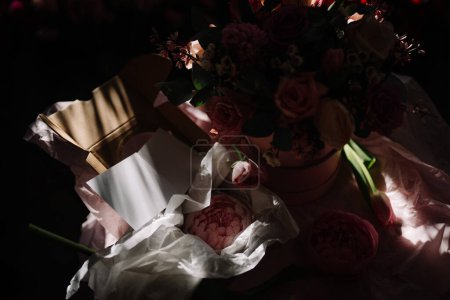 Valmiera, Letonia - 7 de marzo de 2024 - Una foto débilmente iluminada que captura una caja de flores con varias flores y una tarjeta envuelta por sombras, creando una atmósfera malhumorada.