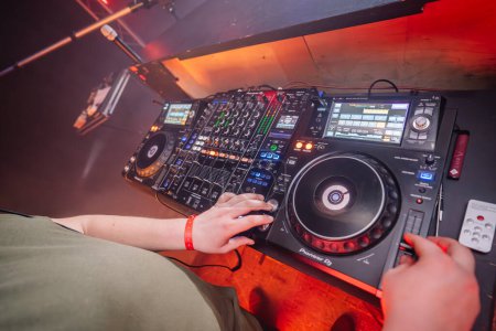 Valmiera, Latvia - March 15, 2024 - Close-up of hands adjusting a DJ mixer and turntables with colorful buttons and screens, set in a club with a reddish light in the background.