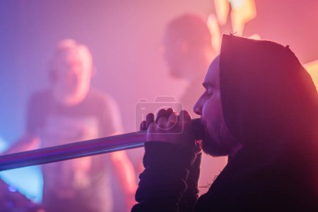 Valmiera, Latvia - March 15, 2024 - A man in a hooded robe plays the didgeridoo, with others in soft focus behind him under pink and blue stage lights.