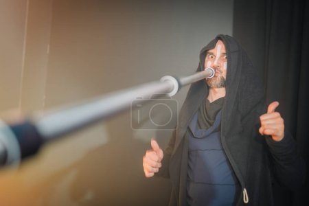 Un hombre con una túnica negra con capucha toca una tuba, mirando a la cámara con una expresión seria, dando un pulgar hacia arriba con un fondo borroso.