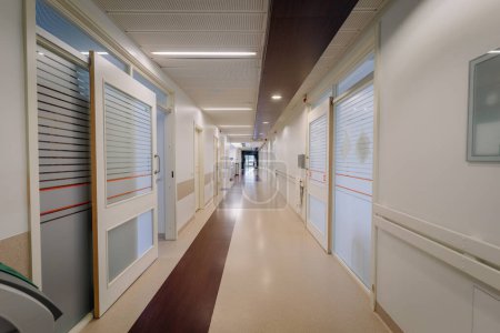 a hospital corridor with doors on either side. The hallway has a shiny floor, ceiling lights, and handrails on the walls.