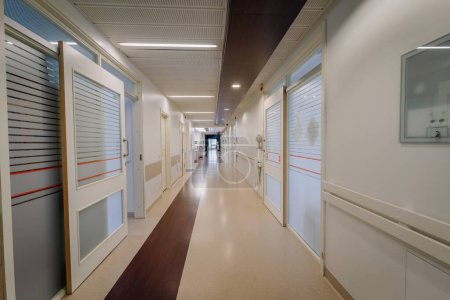 un pasillo del hospital con puertas a cada lado. El pasillo tiene un piso brillante, luces de techo y pasamanos en las paredes.