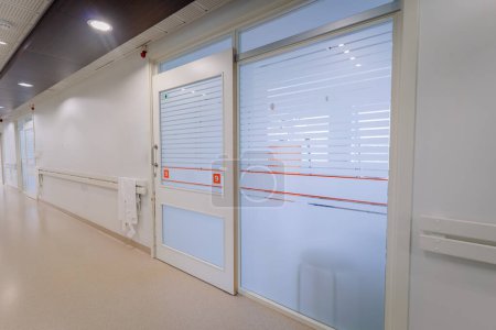 a bright hospital corridor with numbered doors, frosted glass windows, handrails, and a paper towel dispenser.