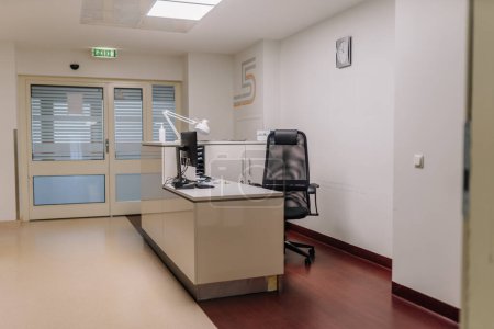 Büroeinrichtung in einem Krankenhaus mit Schreibtisch, Computer, Bürostuhl und Lampe, Nummer 5 an der Wand und Uhr mit Uhrzeit.
