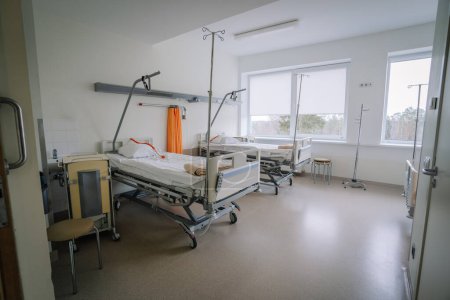 Valmiera, Lettonie - 20 mars 2024 - Une chambre d'hôpital avec deux lits vides, des stands IV, une chaise, de grandes fenêtres avec vue et divers équipements médicaux.