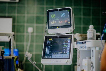 Foto de Valmiera, Letonia - 20 de marzo de 2024 - Las pantallas de la máquina de anestesia con signos vitales para el paciente, colocadas en una sala quirúrgica de azulejos con equipo médico y parte de un proveedor de atención médica visible. - Imagen libre de derechos