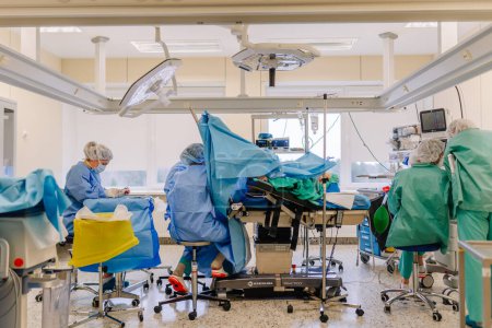Téléchargez les photos : Valmiera, Lettonie - 20 mars 2024 - une équipe chirurgicale se concentre sur une intervention chirurgicale dans une salle d'opération avec un patient sur la table d'opération, drapé de draps bleus stériles, avec des moniteurs médicaux et de l'équipement autour. - en image libre de droit