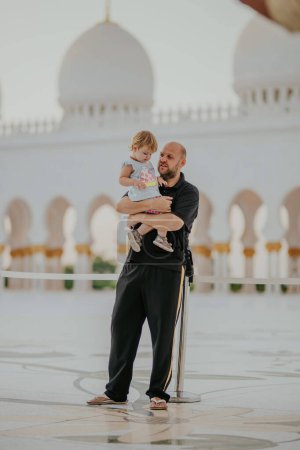 Dubai,  Unite dArab Emirates - October 19, 2019 - Father holding his young child in front of a mosque, both looking off to the side thoughtfully.