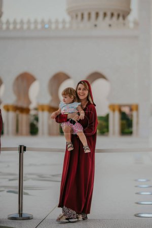 Dubai, Vereinigte Arabische Emirate - 19. Oktober 2019 - Eine lächelnde Frau in einem roten Kleid hält ein Kind vor der weißen Arkade einer Moschee.