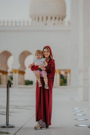 Dubai,  Unite dArab Emirates - October 19, 2019 - A smiling woman in a red dress holding a child, standing in front of a mosque's white arcade.