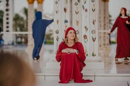 Dubai, Emiratos Árabes Unidos - 19 de octubre de 2019 - Mujer sentada en rojo con pañuelo en la cabeza, mirando hacia arriba, pilar adornado y figuras en vestido tradicional detrás de ella.