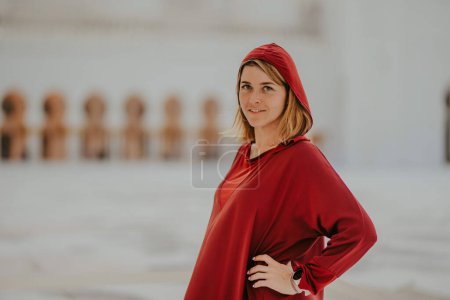 Dubai,  Unite dArab Emirates - October 19, 2019 - Woman in a red tunic and headscarf touching her hair, with a mosques blurred arcade in the background