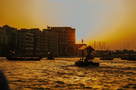 Dubai, Vereinigte Arabische Emirate - 19. Oktober 2019 - Eine heitere Flussszene bei Sonnenuntergang mit Booten und städtischen Gebäuden im Hintergrund, die in goldenes Licht getaucht sind.