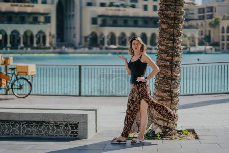 Dubai, Vereinigte Arabische Emirate - 19. Oktober 2019 - Eine Frau in schwarzem Tank-Top und gemustertem Rock steht auf einer Promenade an einer Palme, im Hintergrund ein Gewässer und Gebäude.