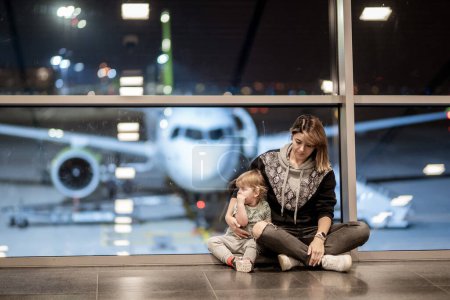 Riga, Lettonie - Le 19 octobre 2019 - Une femme et un tout-petit sont assis par terre près d'une fenêtre de l'aéroport, regardant un avion stationné dehors la nuit.