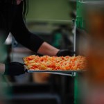 A person in black gloves is handling a tray of orange freeze-dried carrot slices in a blurred kitchen setting.