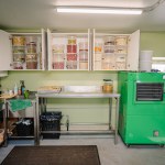 A well-organized garage space with a large green freeze-dryer, metal worktable, and cabinets filled with jars of freeze-dried fruits and supplies.
