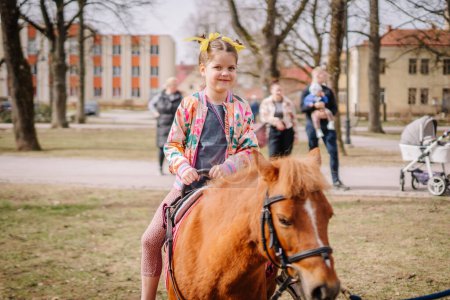 Valmiera, Lettland, 1. April. 2024 - Ein lächelndes Kind auf einem Pony hält die Zügel in der Hand, trägt eine bunte Jacke, mit Schaulustigen und einer Parkanlage im Hintergrund.