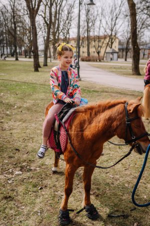 Valmiera, Lettland, 1. April. 2024 - Ein fröhliches Mädchen reitet in einem Park auf einem Pony, trägt bunte Kleidung und hält die Zügel in der Hand, mit Bäumen und einem Pfad im Hintergrund.