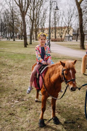 Valmiera, Lettonie, le 1er avril. 2024 - Une fille joyeuse monte un poney dans un parc, portant des vêtements colorés et tenant les rênes, avec des arbres et un chemin en arrière-plan.