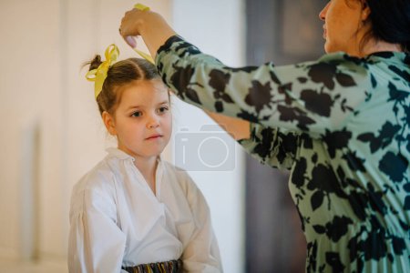 Valmiera, Lettland, 1. April. 2024 - Ein Erwachsener passt ein gelbes Haarband an ein junges Mädchen in weißer Bluse an und bereitet sich auf einen lettischen Volkstanz vor..