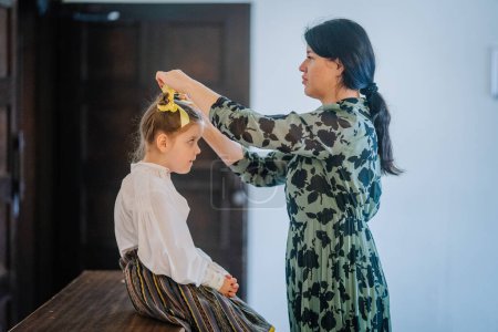 Valmiera, Lettland, 1. April. 2024 - Eine Frau bindet ein gelbes Haarband um ein konzentriertes kleines Mädchen in traditioneller lettischer Kleidung und bereitet sich auf einen Volkstanz vor..