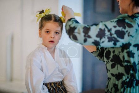 Valmiera, Lettland, 1. April. 2024 - Ein Erwachsener passt ein gelbes Haarband an ein junges Mädchen in weißer Bluse an und bereitet sich auf einen lettischen Volkstanz vor..