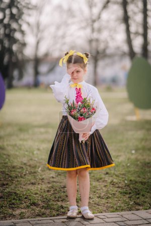 Valmiera, Lettland, 1. April. 2024 - Ein Kind in traditionellem Outfit mit gelben Bändern im Haar wischt eine Träne weg, während es einen Strauß hält.