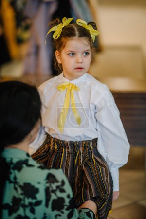 Valmiera, Letonia, 1 de abril. 2024 - Una joven en blusa blanca con lazo amarillo mira hacia otro lado, con una figura adulta borrosa en primer plano.