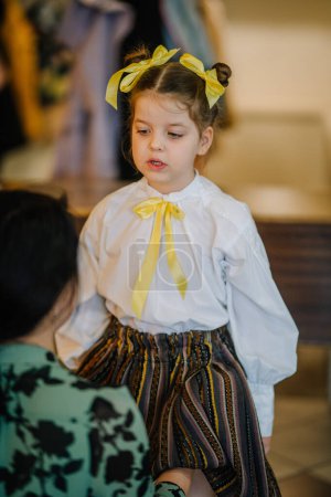 Valmiera, Lettland, 1. April. 2024 - Ein kleines Mädchen in traditioneller lettischer Kleidung blickt zur Seite, mit gelben Bändern im Haar und einer Schleife an der Bluse.