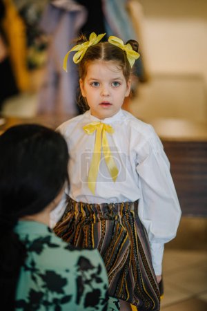 Valmiera, Latvia, April 1. 2024 - A young girl in a white blouse with a yellow bow looks away, with a blurred adult figure in the foreground.