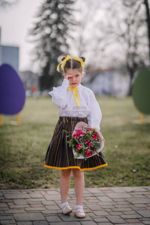 Valmiera, Lettland, 1. April. 2024 - Ein Kind in traditionellem Outfit mit gelben Bändern im Haar wischt eine Träne weg, während es einen Strauß hält.