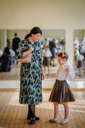Valmiera, Latvia, April 1. 2024 - A woman in a floral dress looks down at a young girl in a traditional skirt and white blouse, both in a room with mirrors.